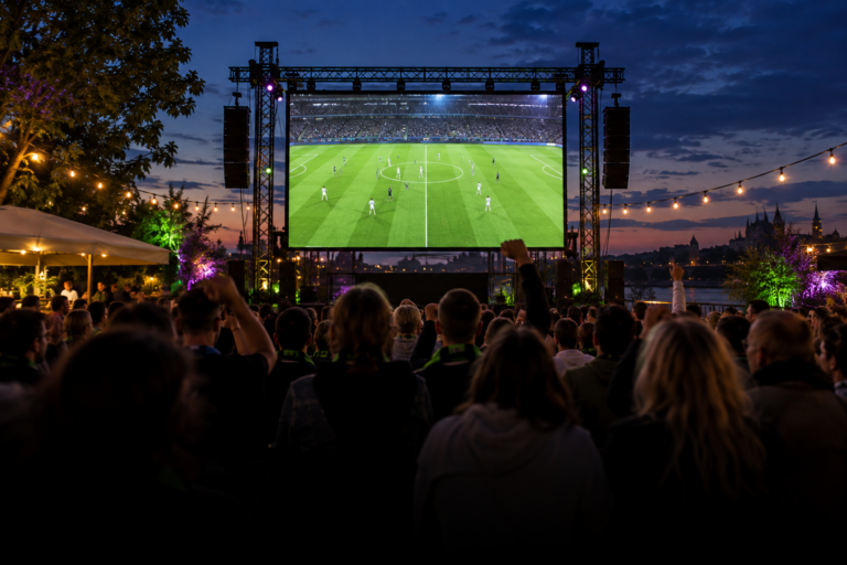 Publikum verfolgt ein Fußballspiel auf einer großen LED-Leinwand unter freiem Himmel in der Abenddämmerung.